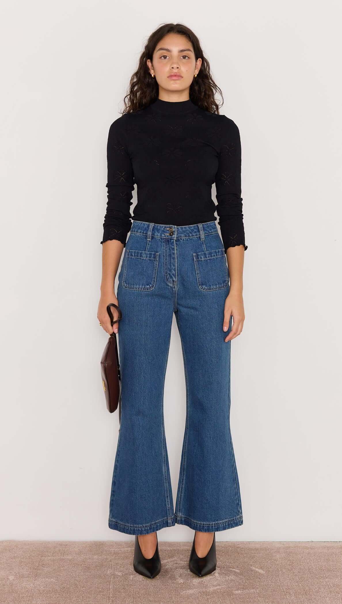 Model wearing high-waisted Nora Bootcut Jeans in vintage blue with a black top and heeled shoes.