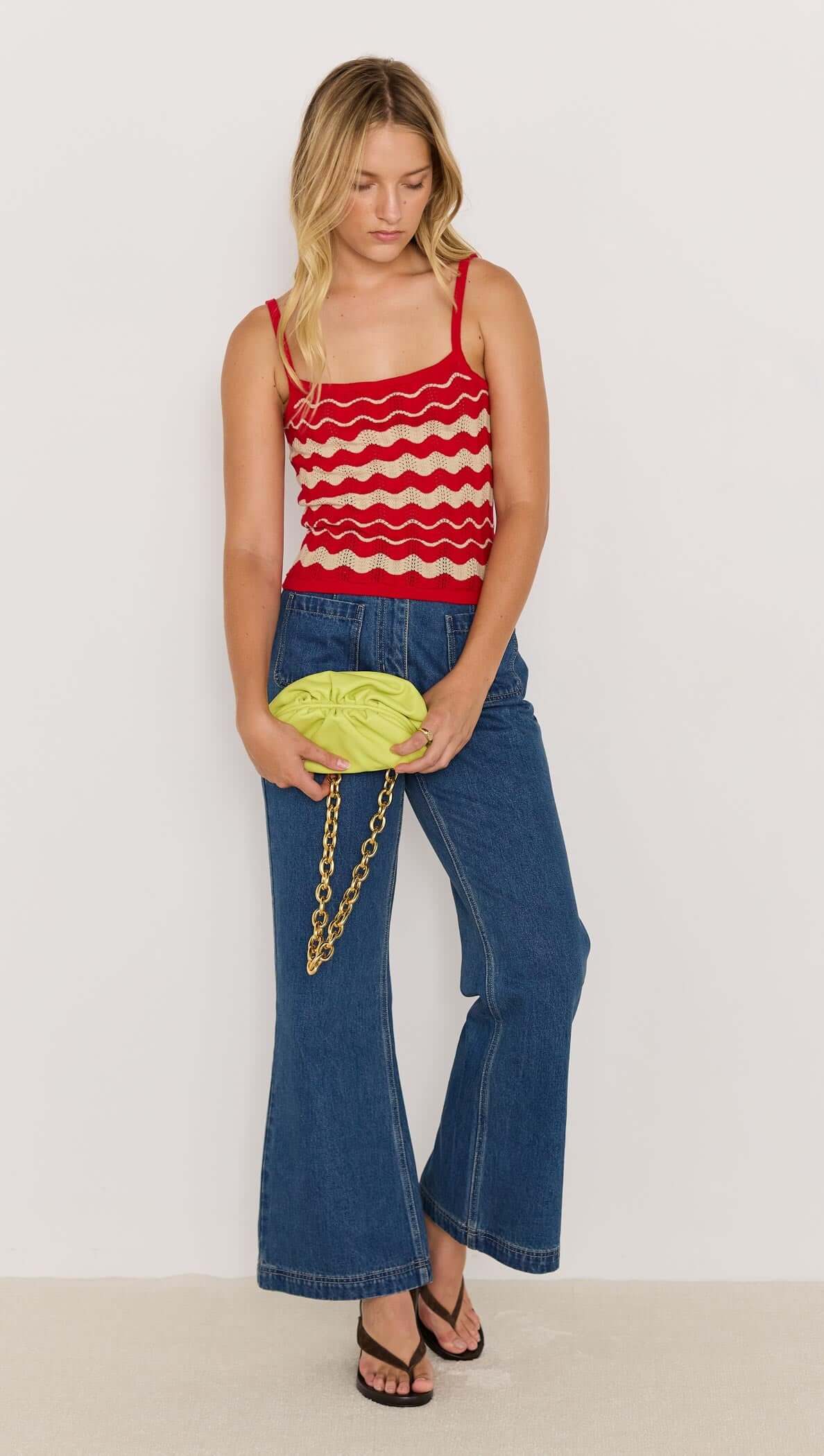 Model wearing a red and beige wave knit cami with denim and a green clutch, showcasing a summer outfit.