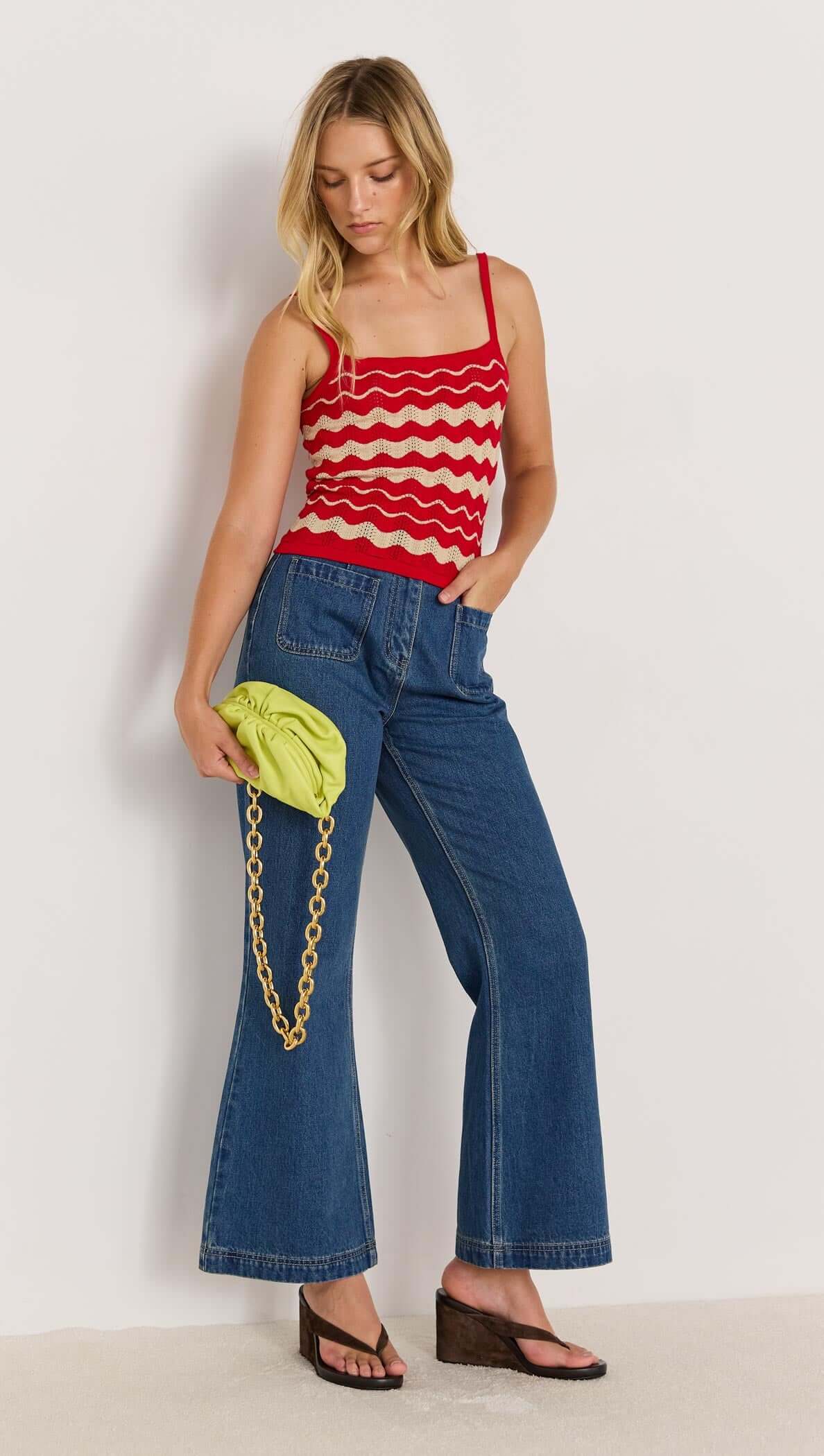 Model wearing a red and beige wave stripe knit cami with denim pants and a green bag, showcasing summer style.