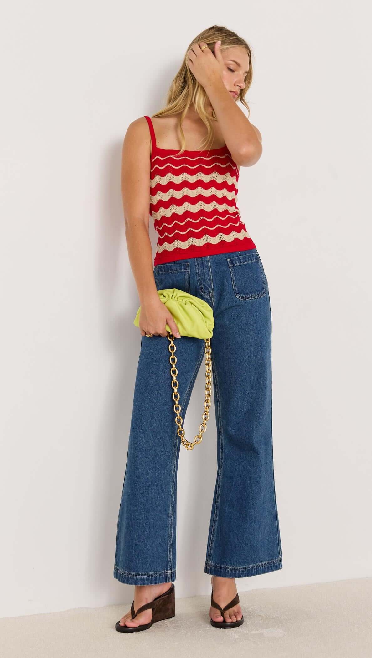 Model wearing a retro-inspired red and beige wave stripe knit cami with denim pants and a neon clutch, showcasing summer fashion.