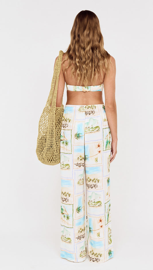 Model wearing Island Postcard Drawstring Beach Pants and a bikini top, holding a patterned tote bag, showcasing a tropical look.