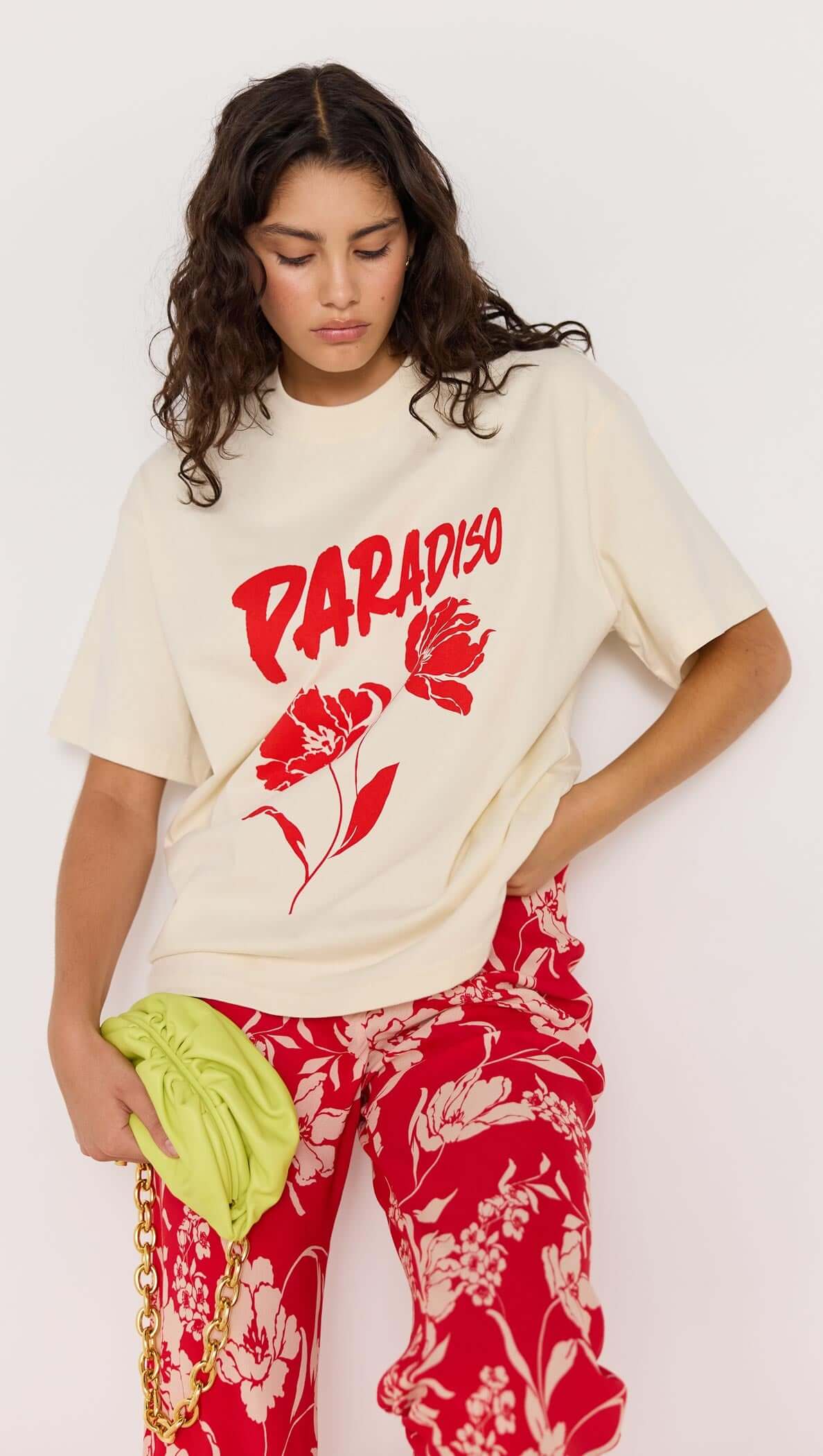 Model wearing cream t-shirt with 'PARADISO' graphic, paired with red floral pants and holding a green clutch.