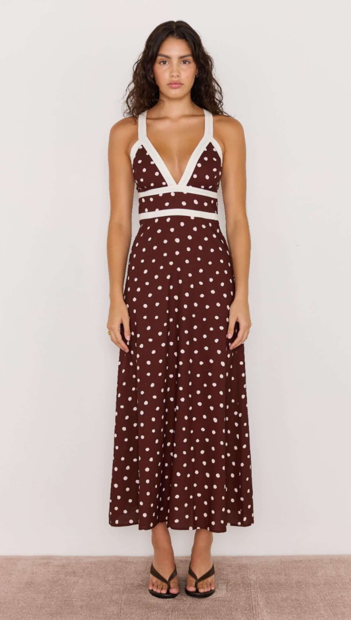 Model wearing a chocolate brown polka dot midi dress with deep V-neckline and cross-back straps, perfect for sunny days.