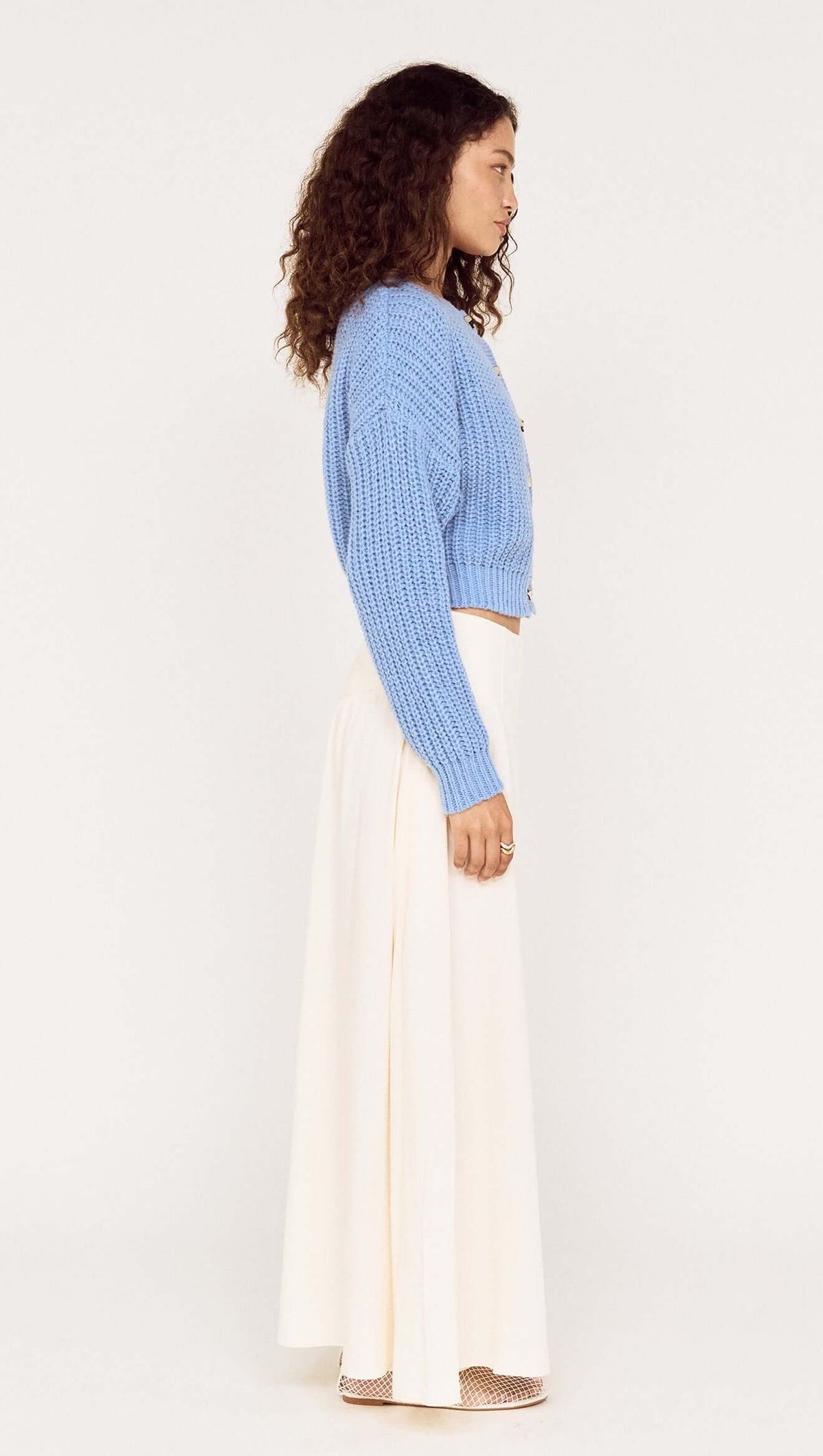 Model wearing a Gold Detail Chunky Knit Cardigan in blue with a cream skirt, showcasing the side view of the outfit.