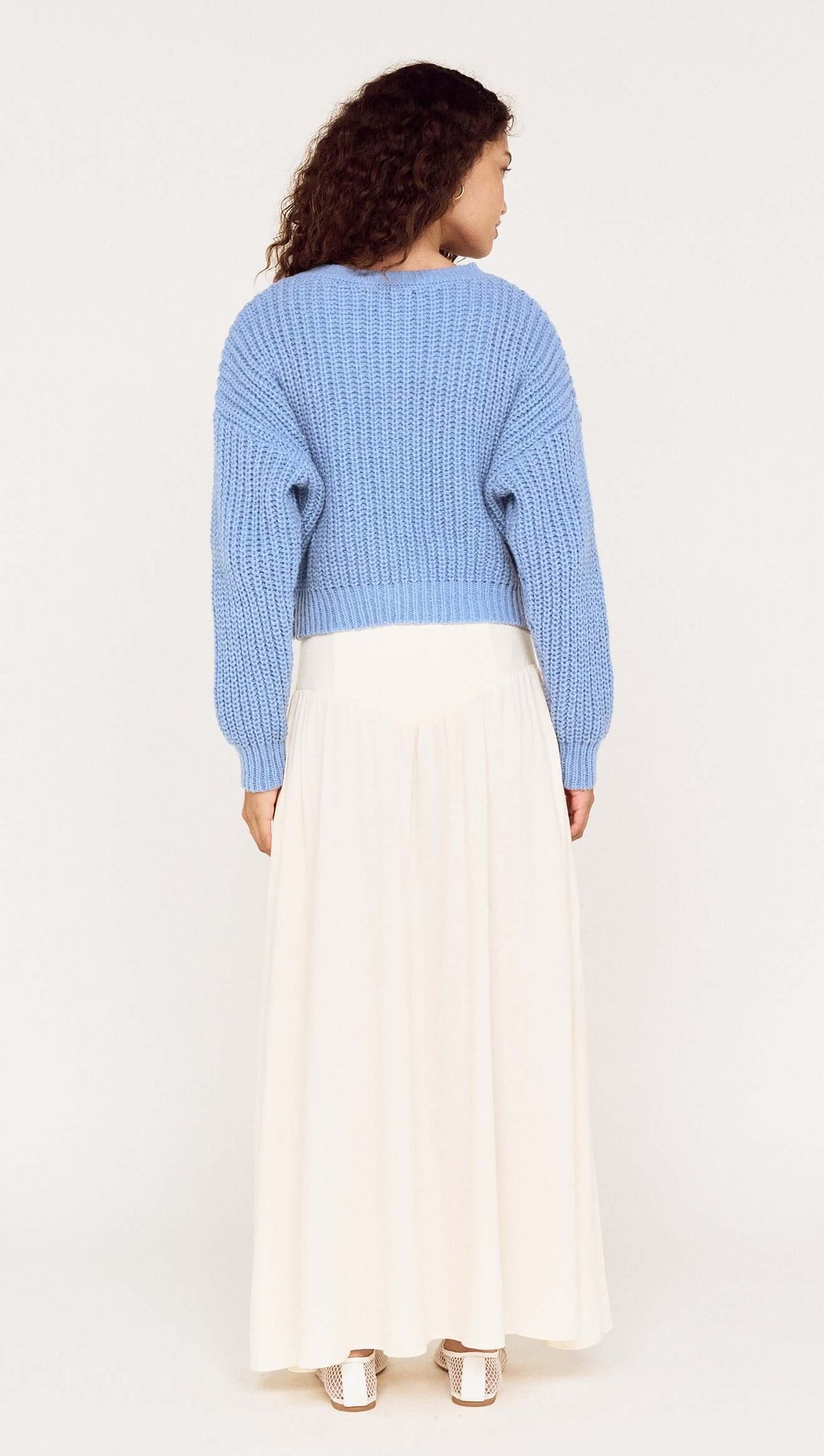 Back view of a model wearing a blue chunky knit cardigan with a long white skirt.