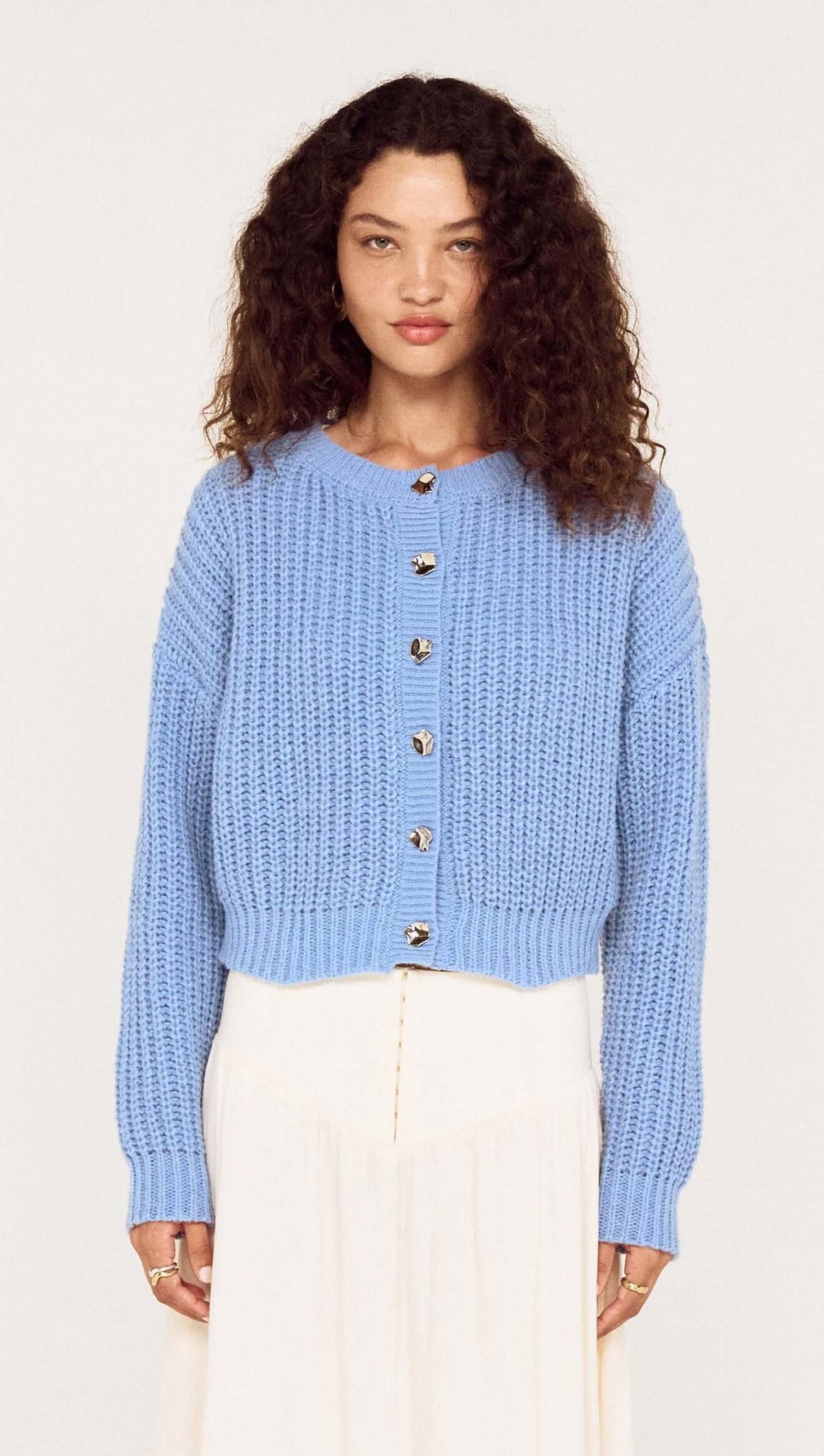 Model wearing a blue chunky knit cardigan with buttons, styled with a flowy white skirt, perfect for layered outfits.