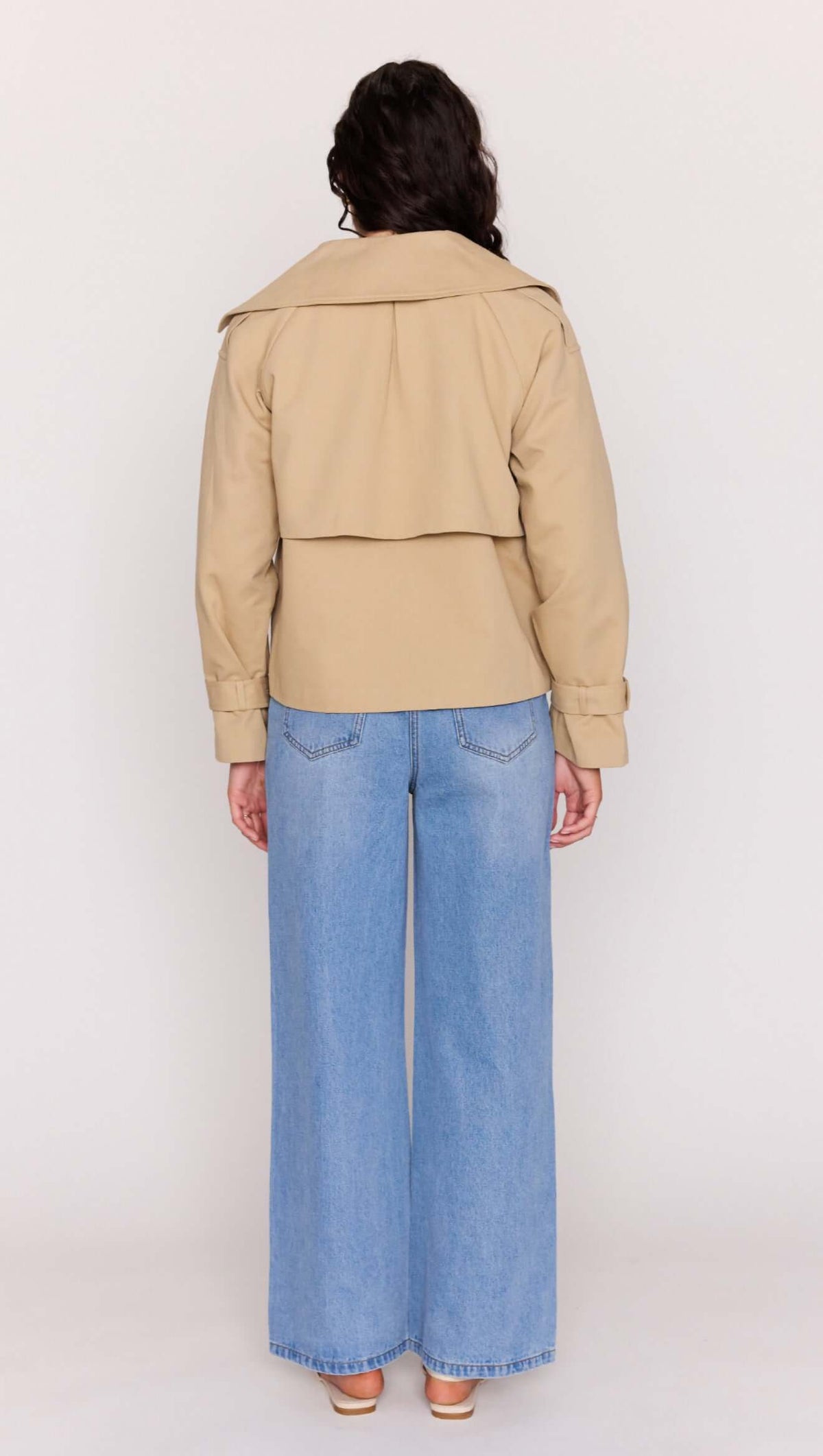 Back view of a woman wearing a Beige Cotton Drill Cropped Trench with wide lapels and loose blue jeans.