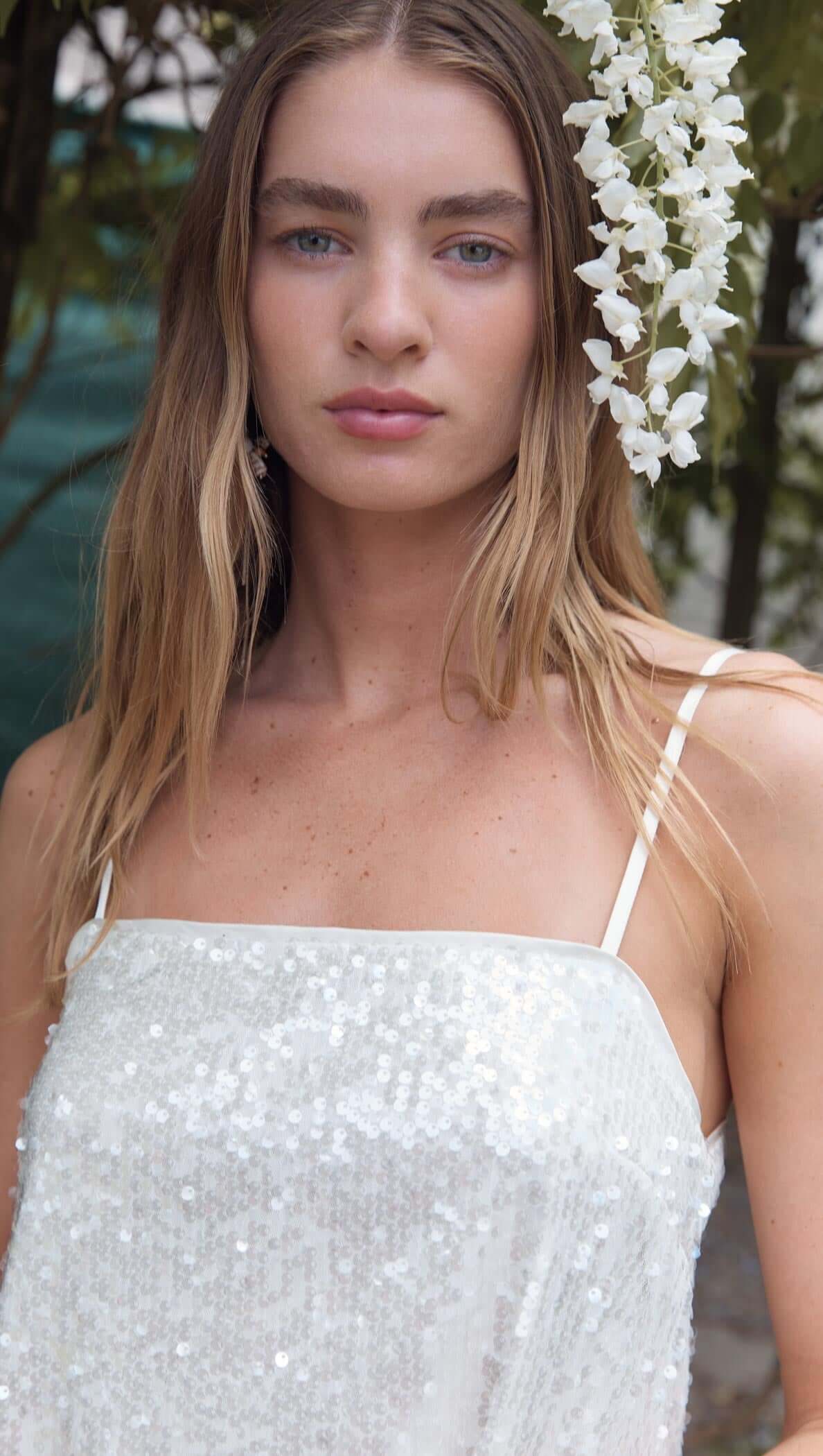 Model wearing a White Sequin Camisole Top with spaghetti straps, adorned with flowers in a beautiful outdoor setting.