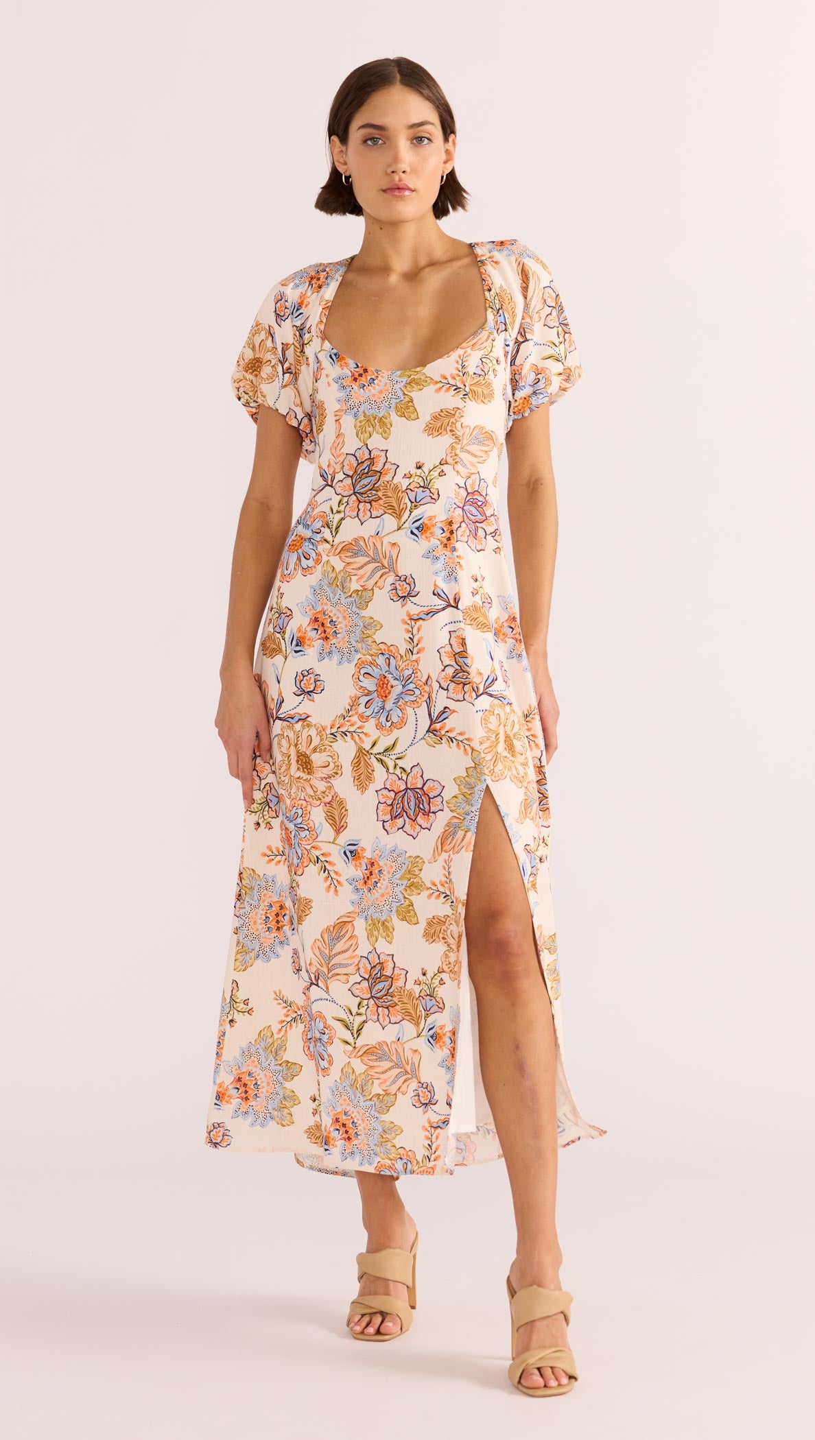 A model wearing a floral midi dress with short puffed sleeves, a sweetheart neckline, and a square back design with a tie closure. The dress features a front slit and is designed with a lightweight fabric.