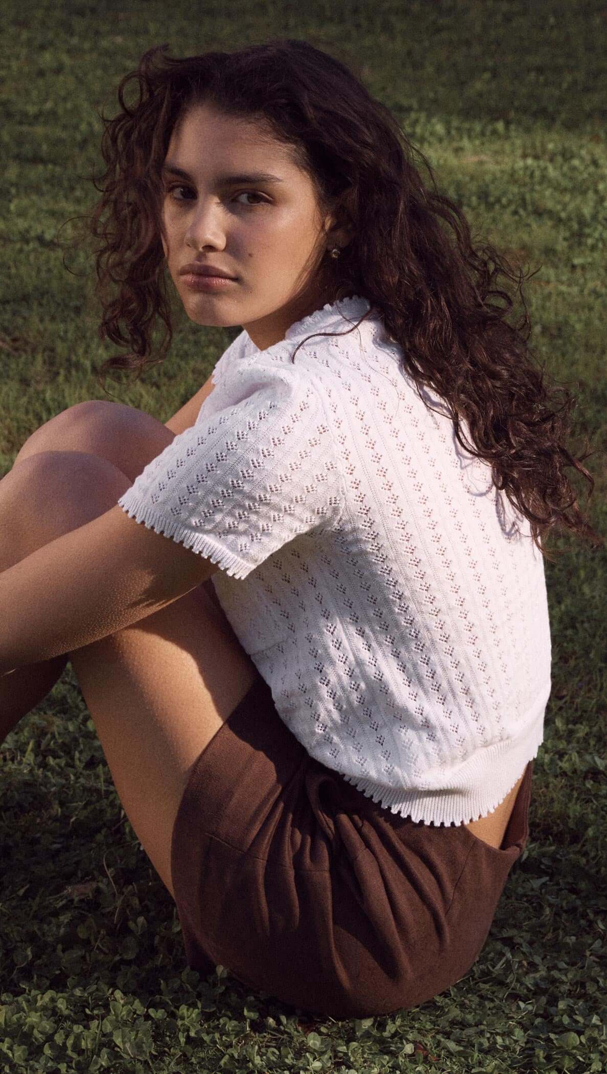Model sitting on grass wearing a white French pointelle short sleeve knit top and brown shorts.