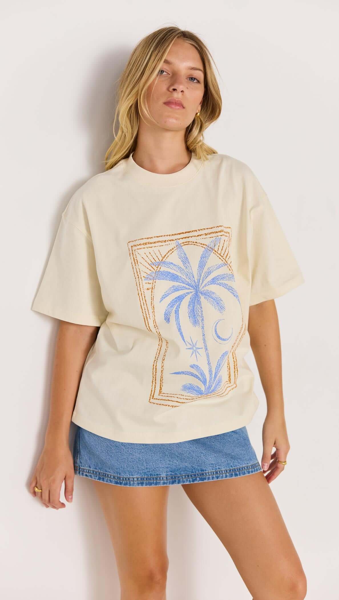 Model wearing a palm print cotton oversize t-shirt styled with a denim skirt in a laid-back coastal setting.