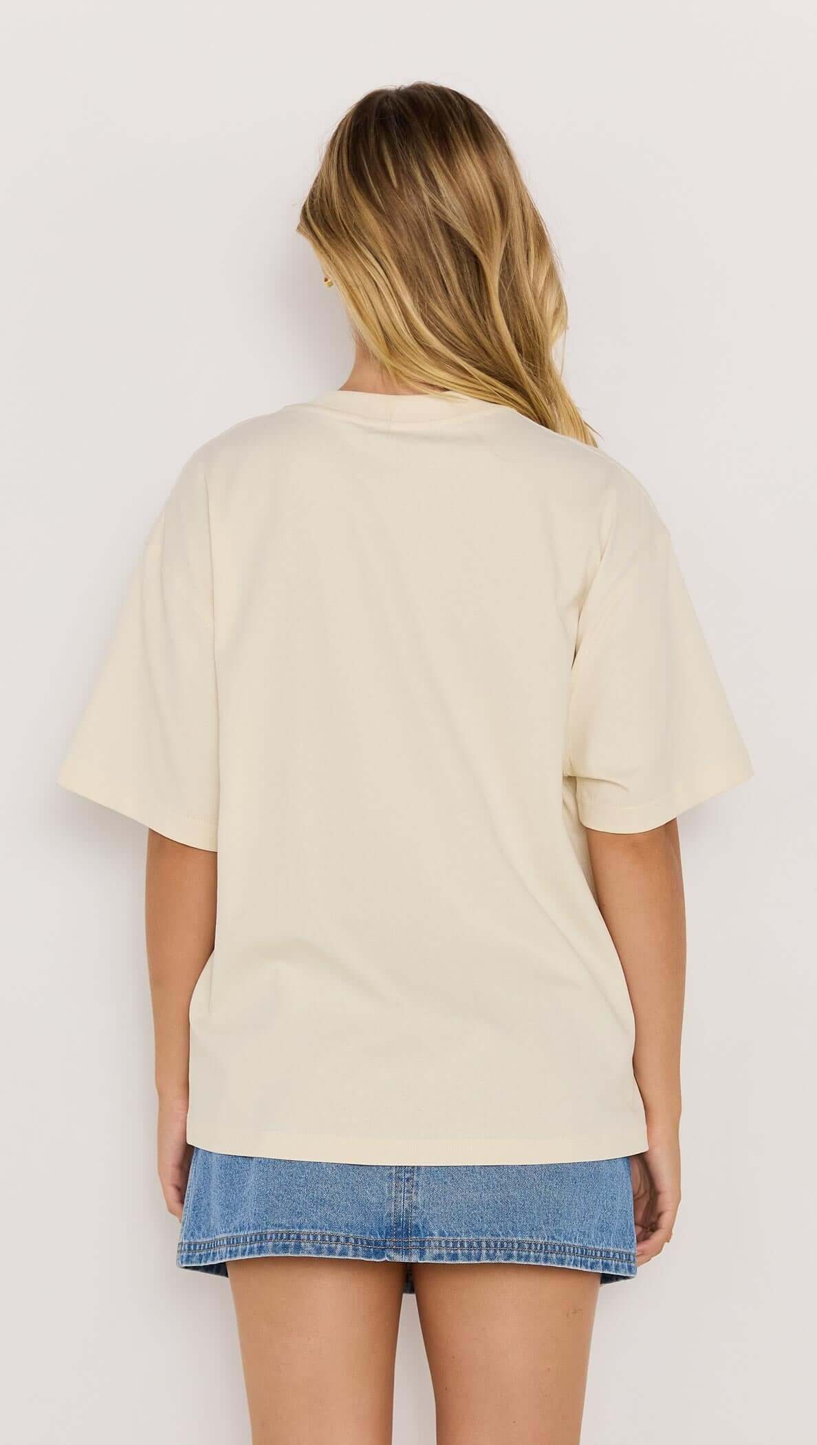 Back view of a woman wearing a beige palm print cotton oversize t-shirt paired with a denim mini skirt.