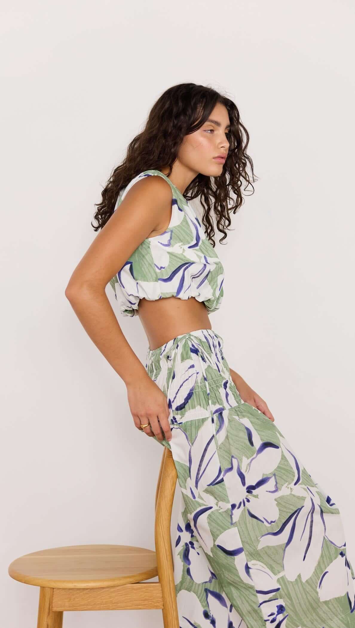 Model wearing green and white floral midi skirt with matching top, posing beside a wooden chair.
