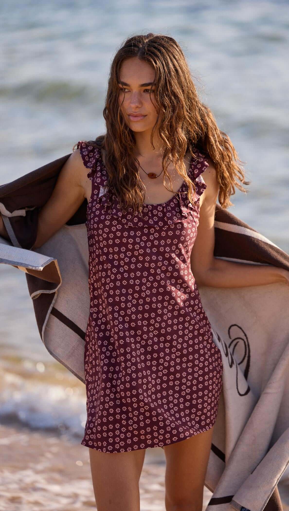 Model wearing a Burgundy Floral Print Ruffle Strap Low-Back Mini Dress at the beach, holding a towel.