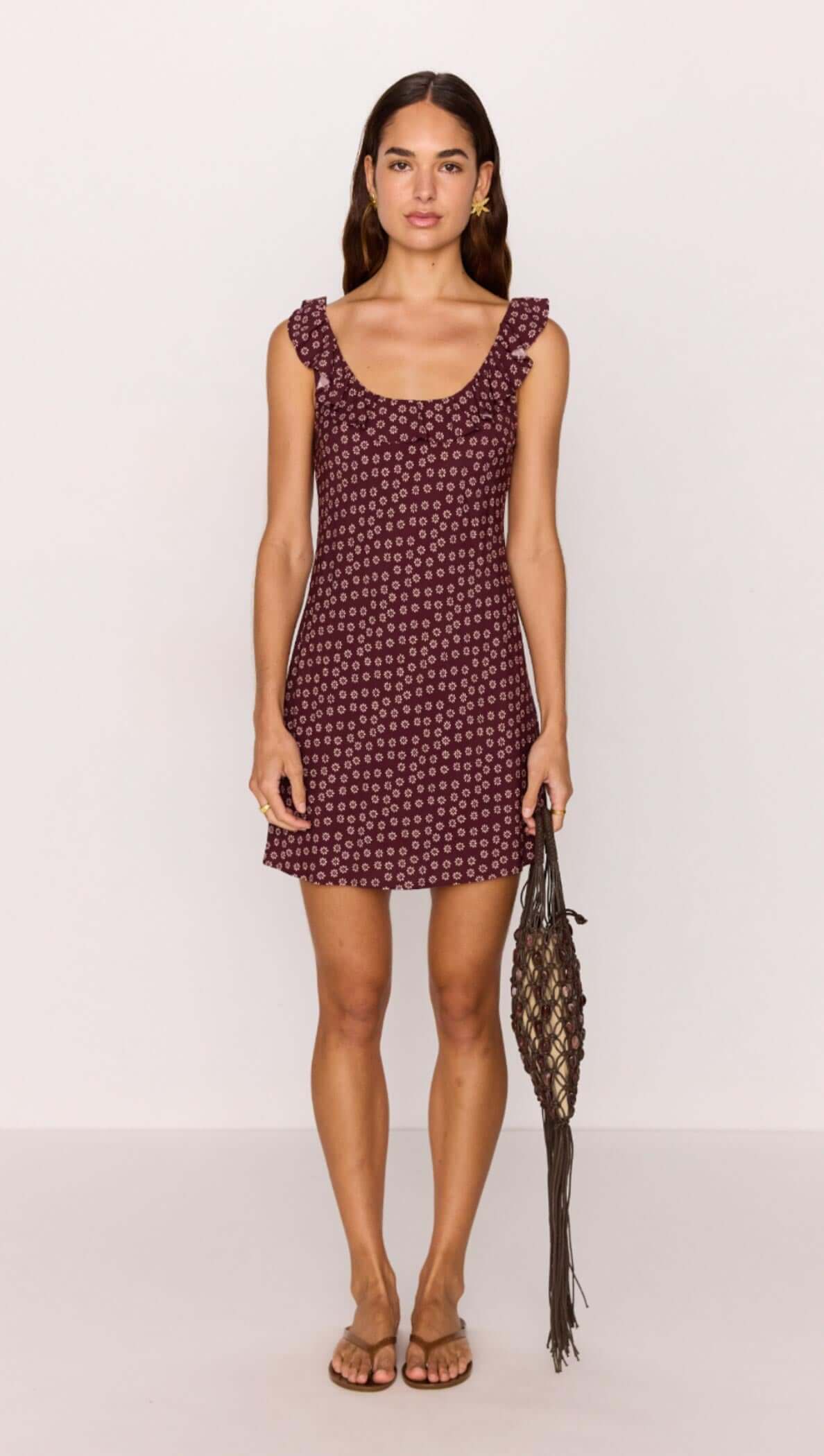 Model wearing a burgundy floral print ruffle strap low-back mini dress, holding a woven bag.