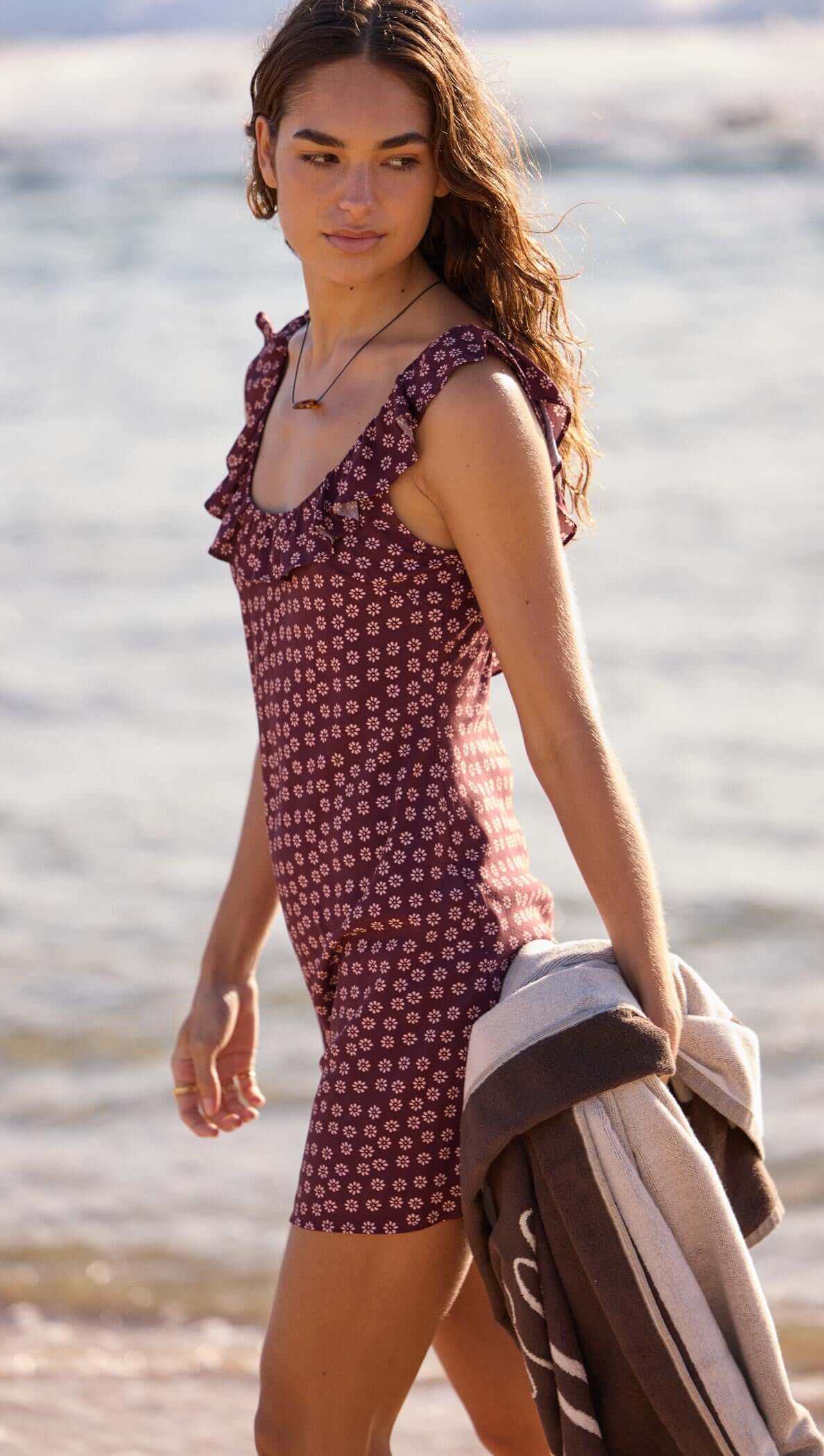 Young woman wearing a ruffle strap burgundy floral mini dress, walking on the beach with a towel.