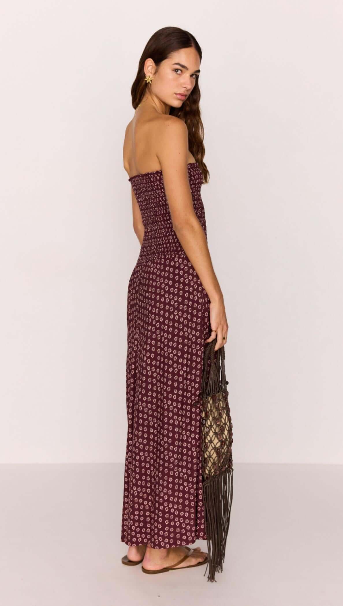 Model wearing a strapless burgundy floral maxi dress, showcasing a ruched bodice and dropped hem, holding a fringed bag.