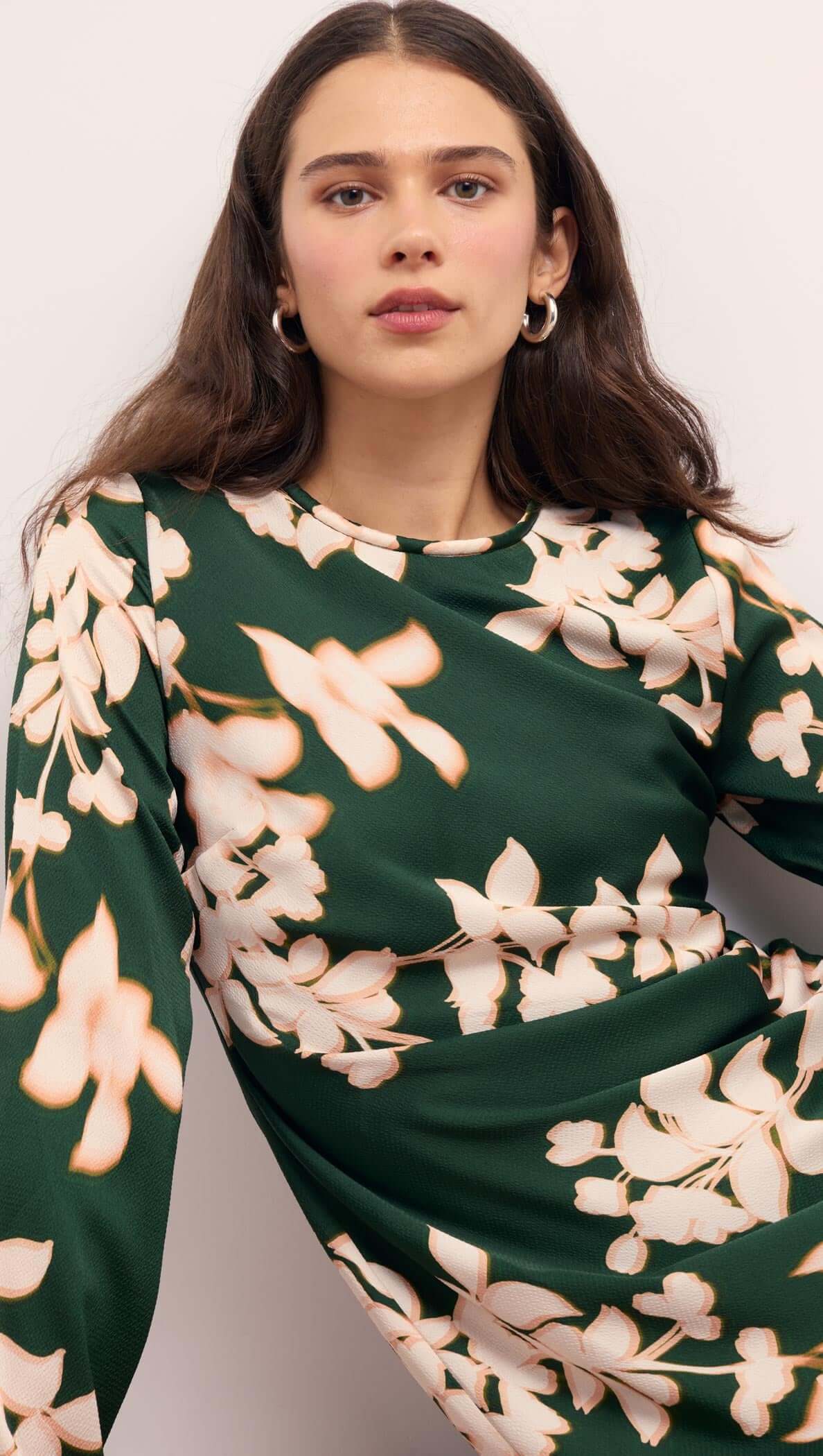 Woman modeling the Green & Cream Floral Satin Mini Dress with a forest green background and floral design accents.