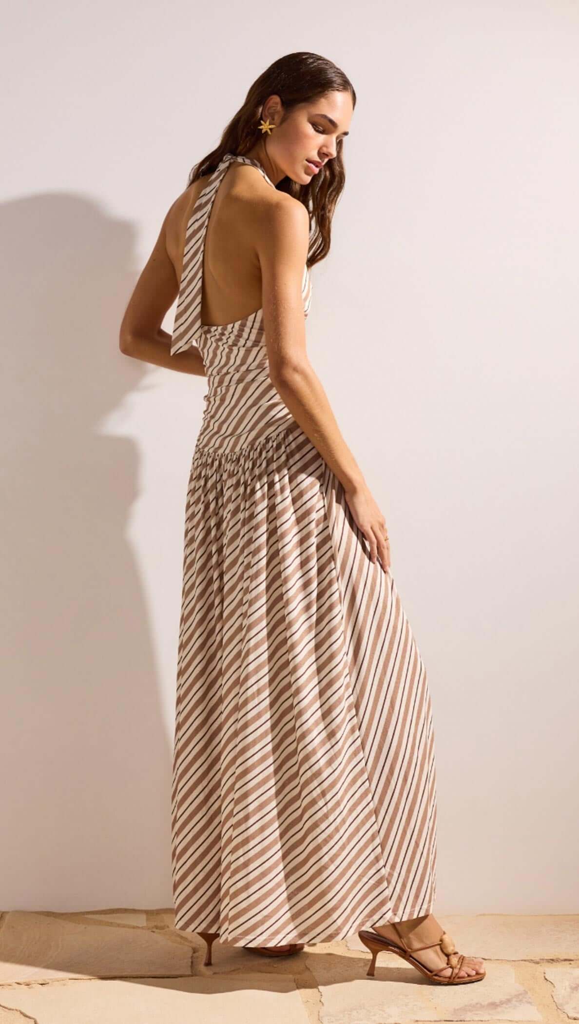 Back view of a model wearing the Brown & Cream Stripe Low-Waist Halter Maxi Dress, showcasing its flowy design and striped pattern.