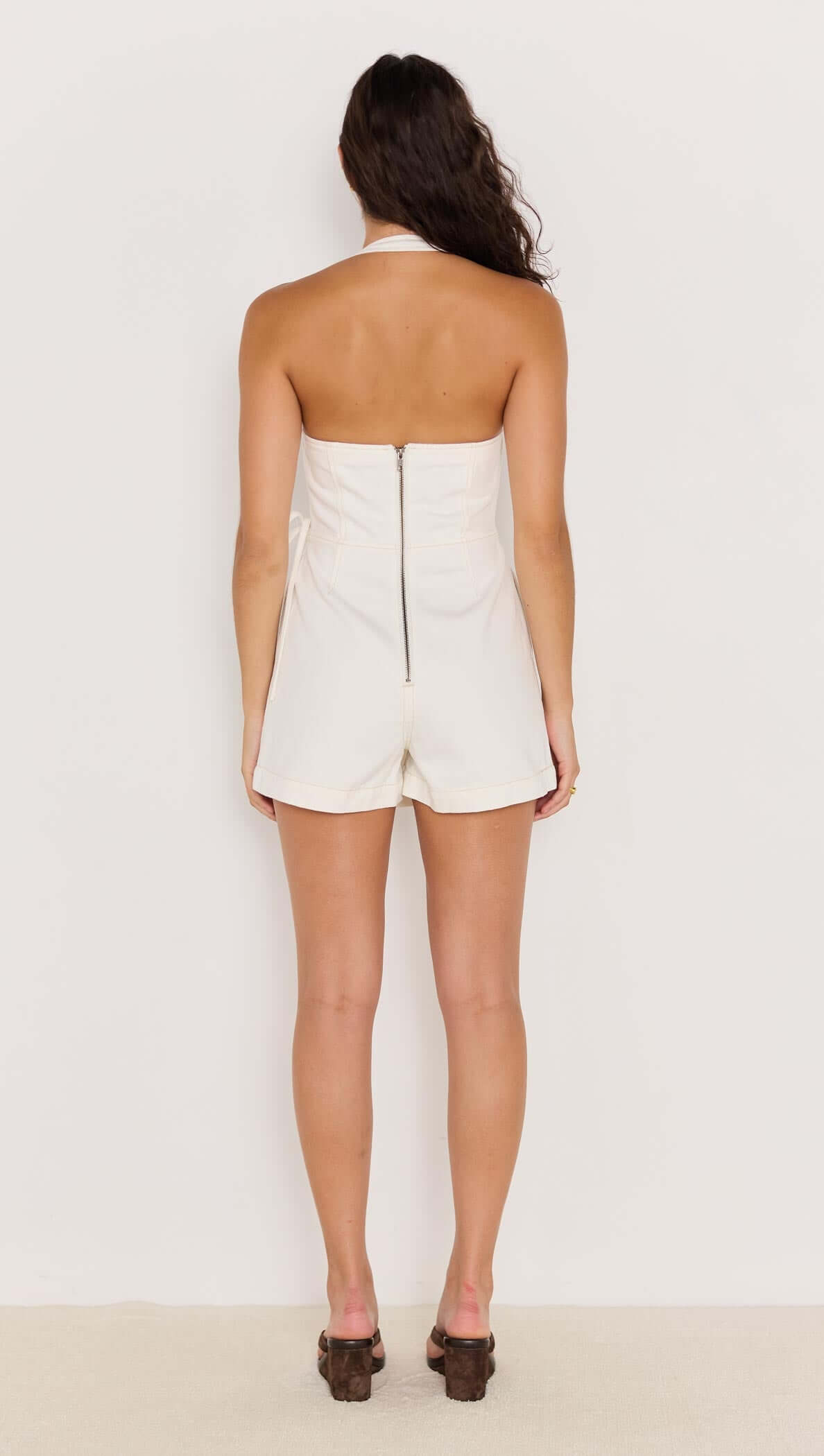 Back view of a woman wearing a white denim wrap halter-neck playsuit with a zipper closure.