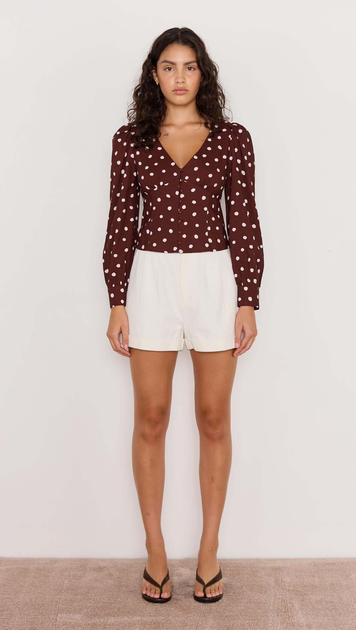 Model wearing the Cedar Spot Corset Blouse in chocolate brown with cream spots, paired with white shorts.