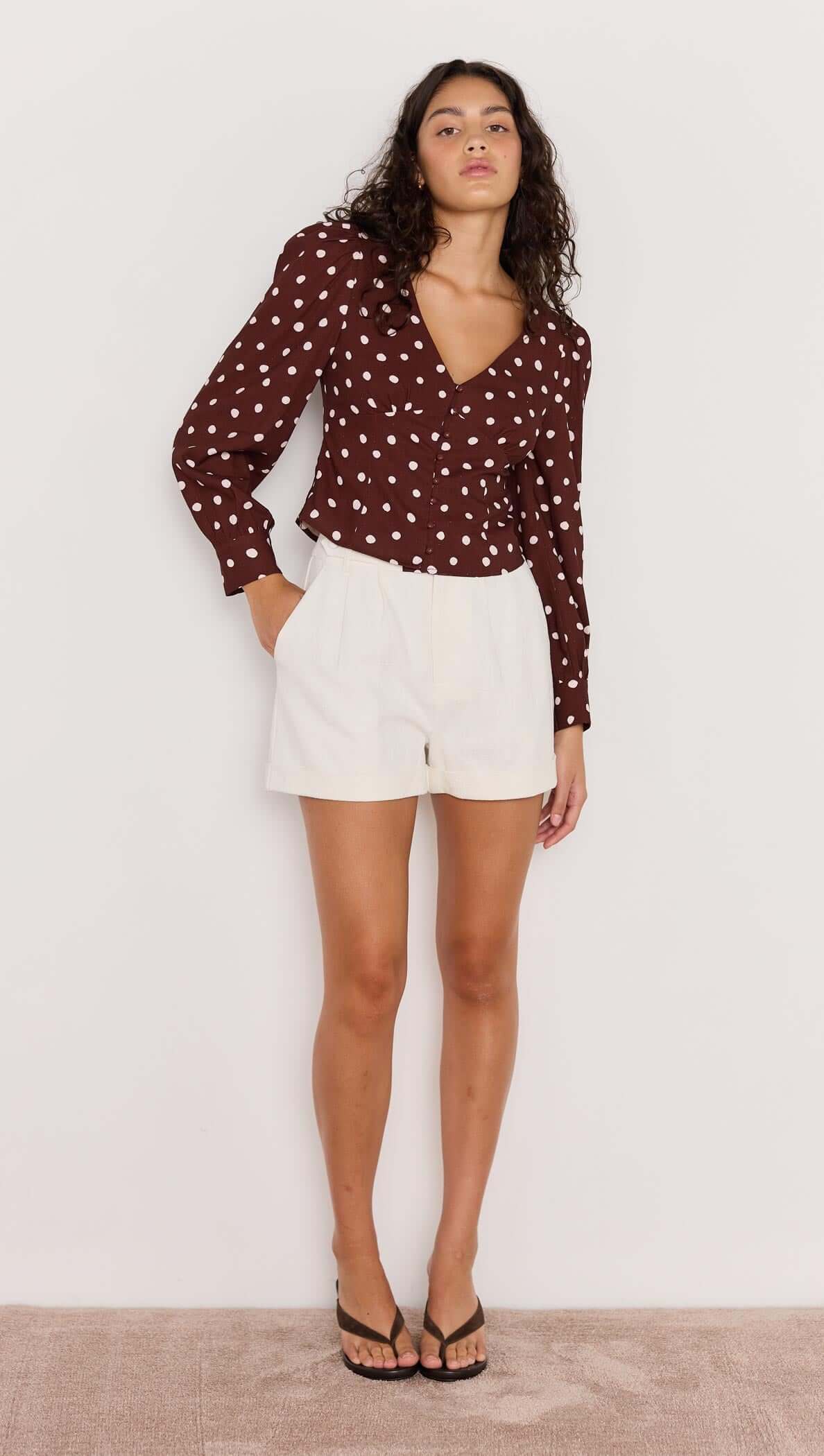 Model wearing the Cedar Corset Blouse in chocolate brown with cream spots, paired with white shorts and flip flops.