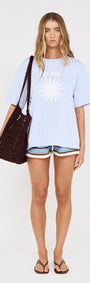 Model wearing Relaxed Fit El Sol Print Tee in Pastel Blue with shorts and a woven bag.