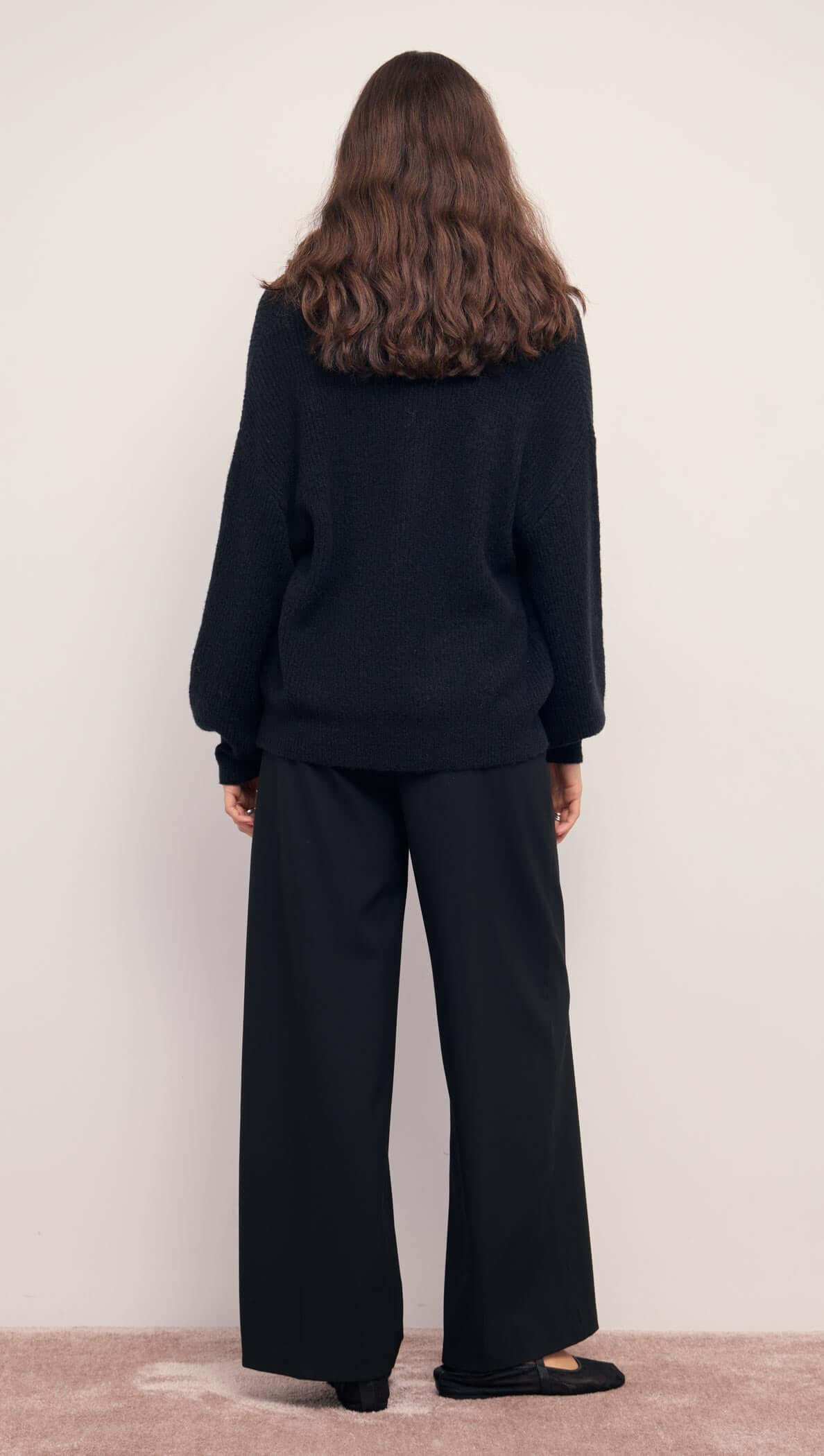 Back view of a model wearing a Relaxed Open Black Cardigan With Gold Closure paired with wide-leg black pants.