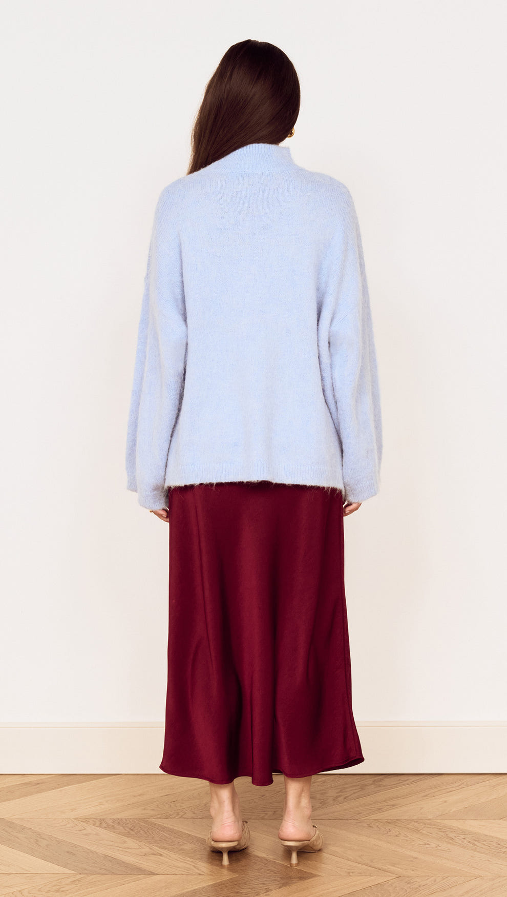 Back view of a model wearing a light blue sweater and a Burgundy Side Split Bias Midi Skirt on a wooden floor.