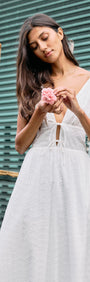 Model wearing a Cotton Broderie Anglaise Sleeveless Dress, holding a flower, with a backdrop of green texture.