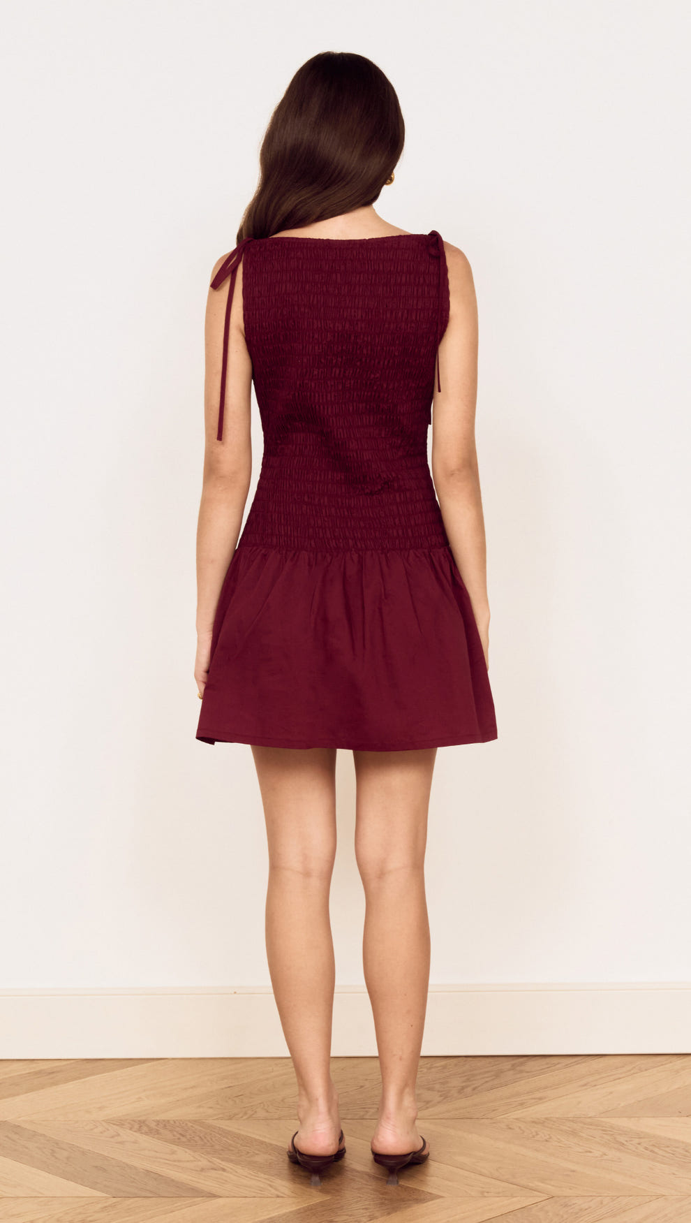 Back view of a woman wearing a Cotton Shirred Mini Dress in Burgundy with tie shoulder details.