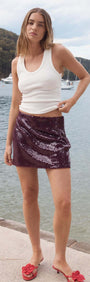 Model wearing a Purple Sequin Mini Skirt by the water, showcasing a stylish party look with a white tank top.
