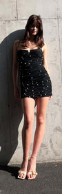 Model wearing Pearl Embellished Black Mesh Mini Dress with pearl accents and ruched finish, posing against a concrete wall.