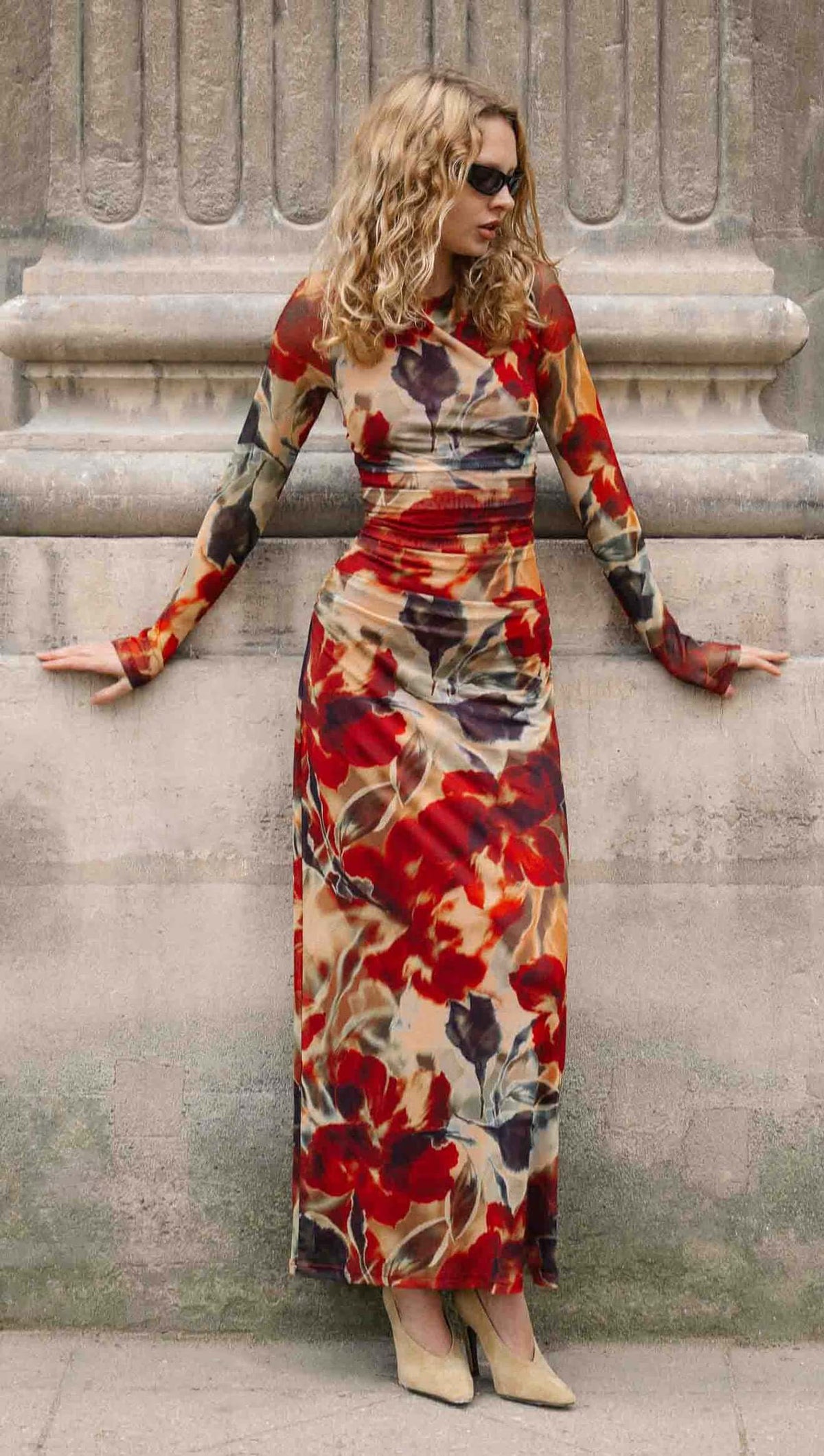 Woman in a Red Floral Sheer Mesh Midi Dress poses against a stone wall, showcasing bold floral patterns and long sleeves.