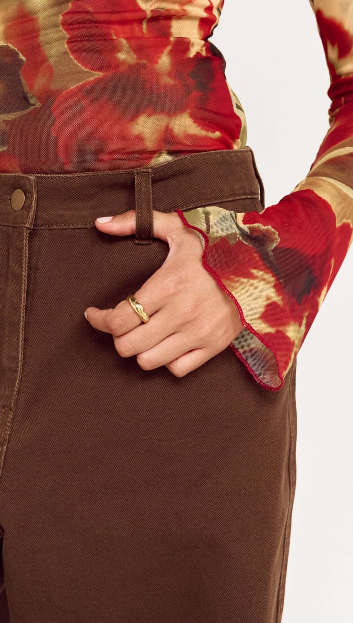 Close-up of a person wearing Brown Mid Rise Barrel Leg Jeans, showing hand on waistband with floral long-sleeve top.