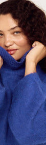 Woman wearing a Blue Oversized Roll Neck Jumper, smiling and holding the collar, showcasing its cozy and stylish design.