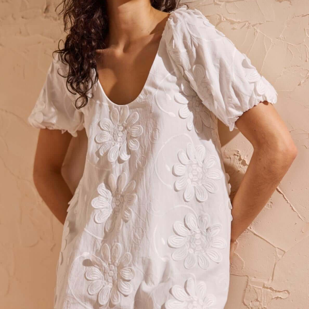 White Cotton Floral Applique Smock Mini Dress on a model with puff sleeves and floral details against a textured background.