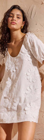 White Cotton Floral Applique Smock Mini Dress on a model with puff sleeves and floral details against a textured background.