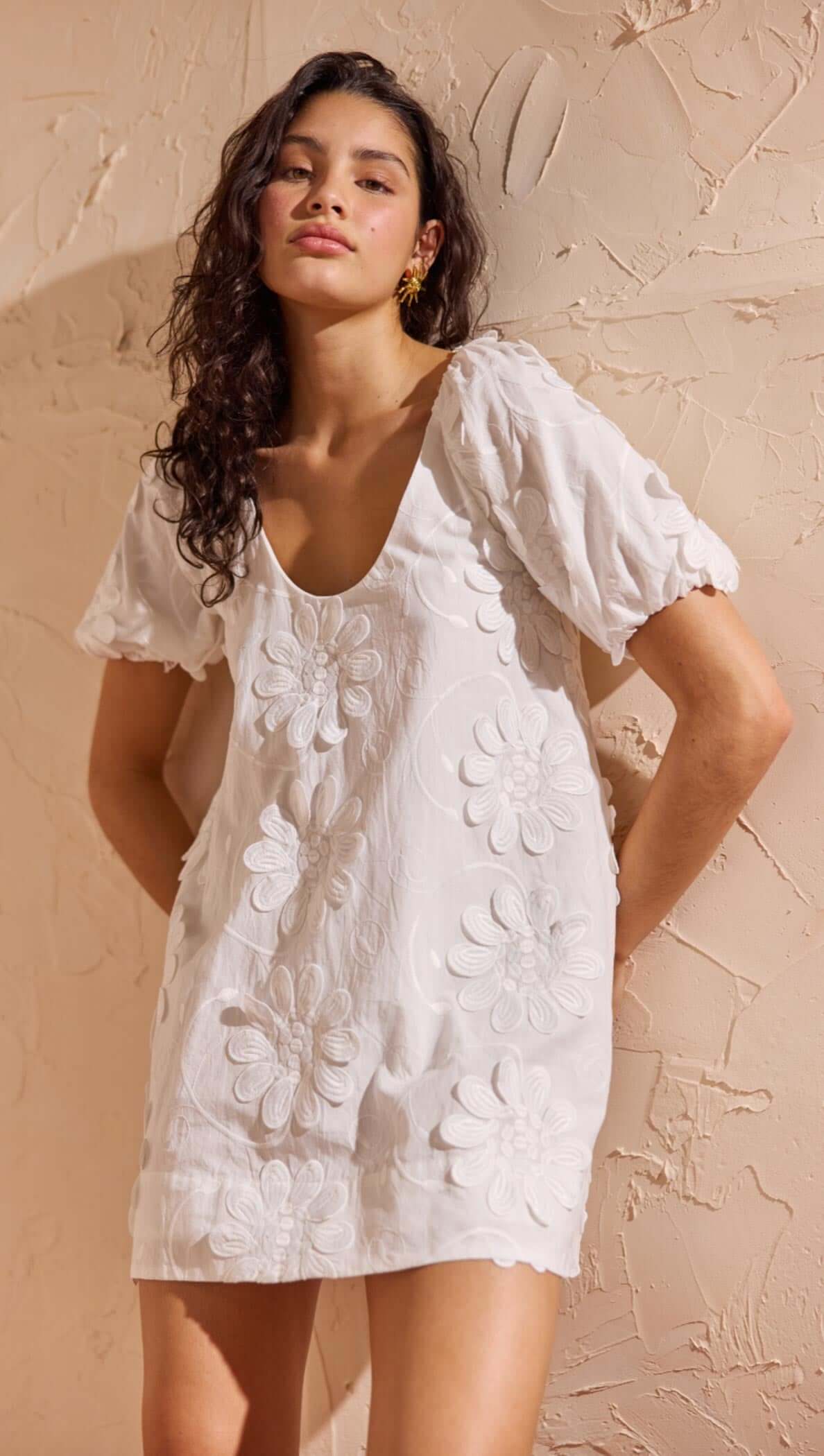 White Cotton Floral Applique Smock Mini Dress on a model with puff sleeves and floral details against a textured background.