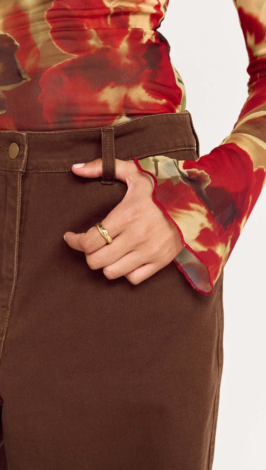 Close-up of a person wearing Brown Mid Rise Barrel Leg Jeans, showing hand on waistband with floral long-sleeve top.