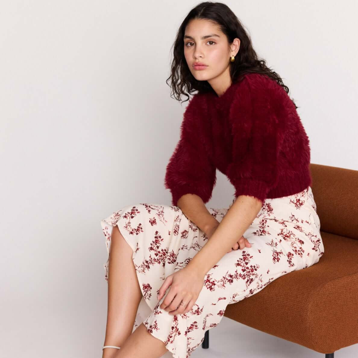 Model wearing a Cream Floral Toile Bias Cut Maxi Skirt paired with a burgundy sweater, seated on a stylish brown chair.