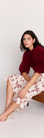 Model wearing a Cream Floral Toile Bias Cut Maxi Skirt paired with a burgundy sweater, seated on a stylish brown chair.
