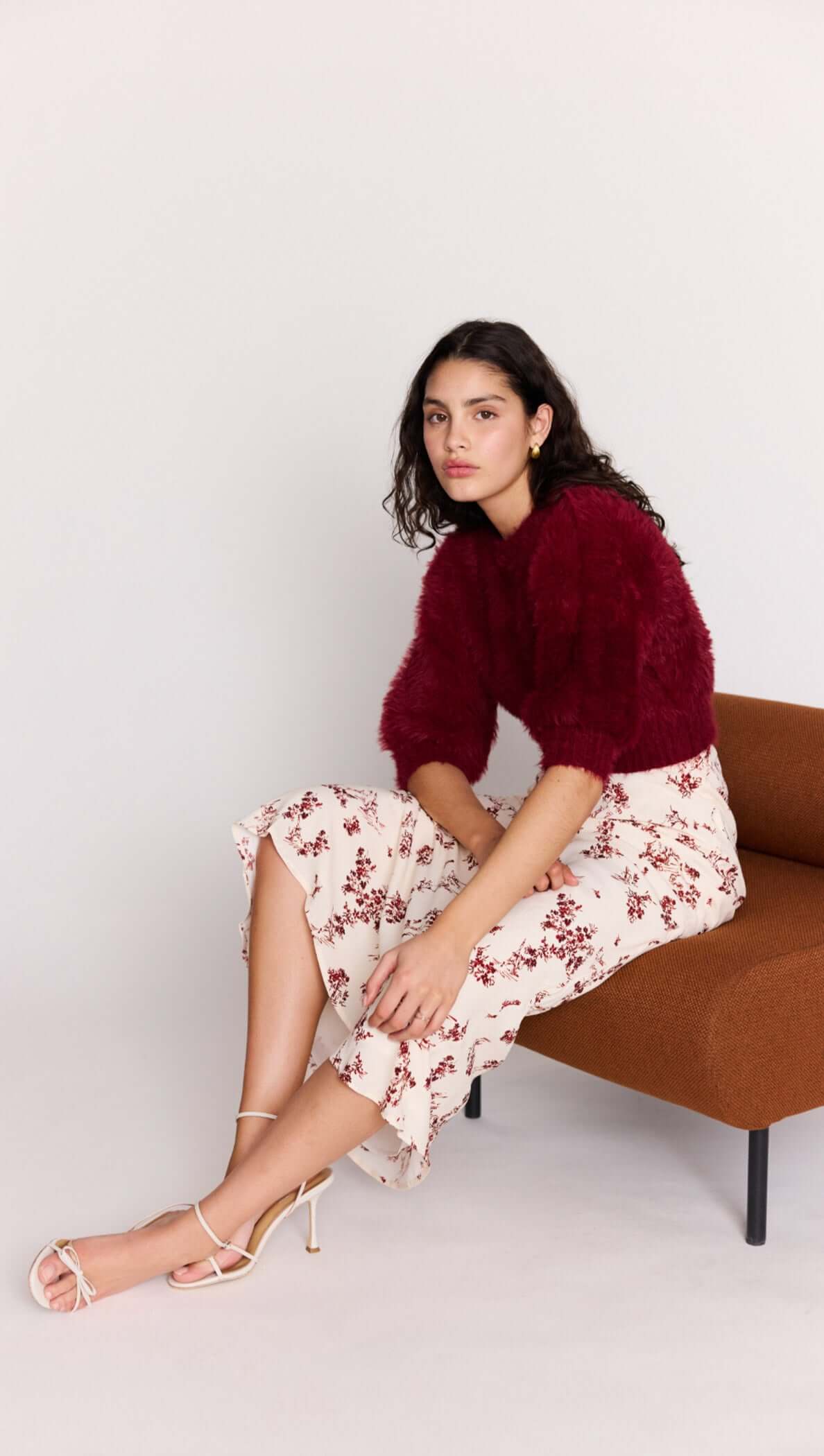 Model wearing a Cream Floral Toile Bias Cut Maxi Skirt paired with a burgundy sweater, seated on a stylish brown chair.
