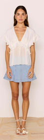Model wearing a Marlon White Ruffle Blouse with flutter sleeves, styled with a denim skirt for a summer look.
