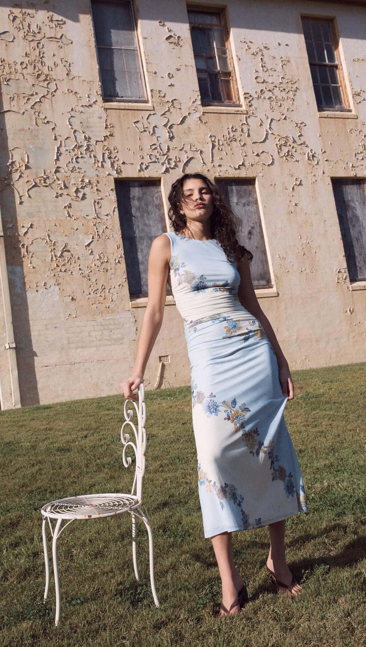 Woman in Lucia blue floral mesh maxi dress against a textured wall, exuding a romantic and effortless style.