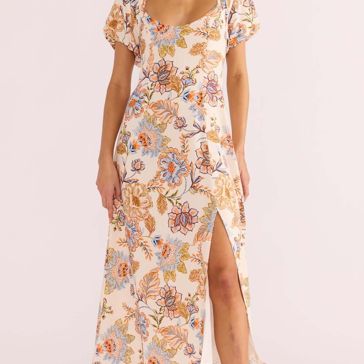 A model wearing a floral midi dress with short puffed sleeves, a sweetheart neckline, and a square back design with a tie closure. The dress features a front slit and is designed with a lightweight fabric.