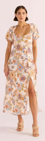 A model wearing a floral midi dress with short puffed sleeves, a sweetheart neckline, and a square back design with a tie closure. The dress features a front slit and is designed with a lightweight fabric.