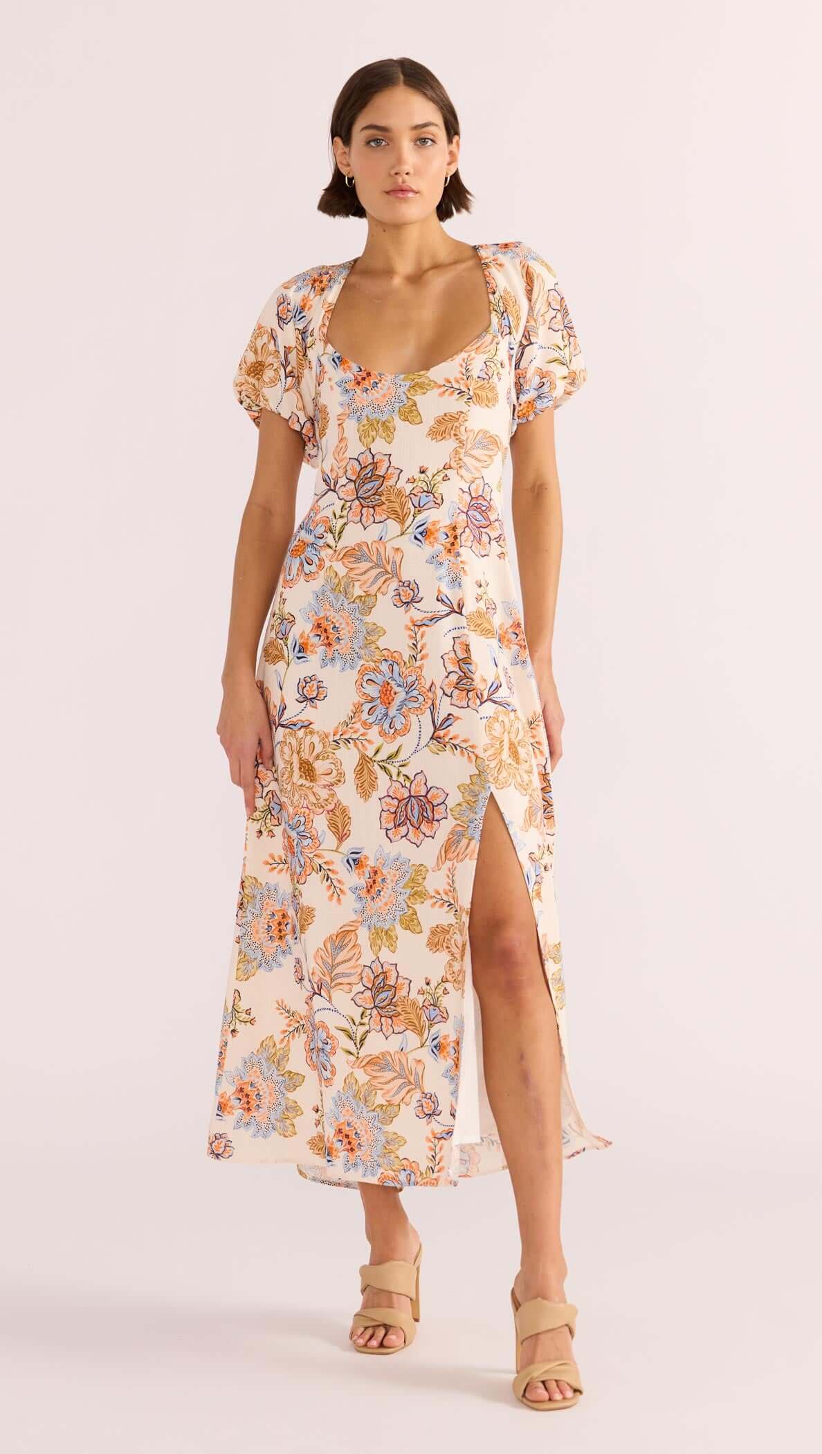 A model wearing a floral midi dress with short puffed sleeves, a sweetheart neckline, and a square back design with a tie closure. The dress features a front slit and is designed with a lightweight fabric.