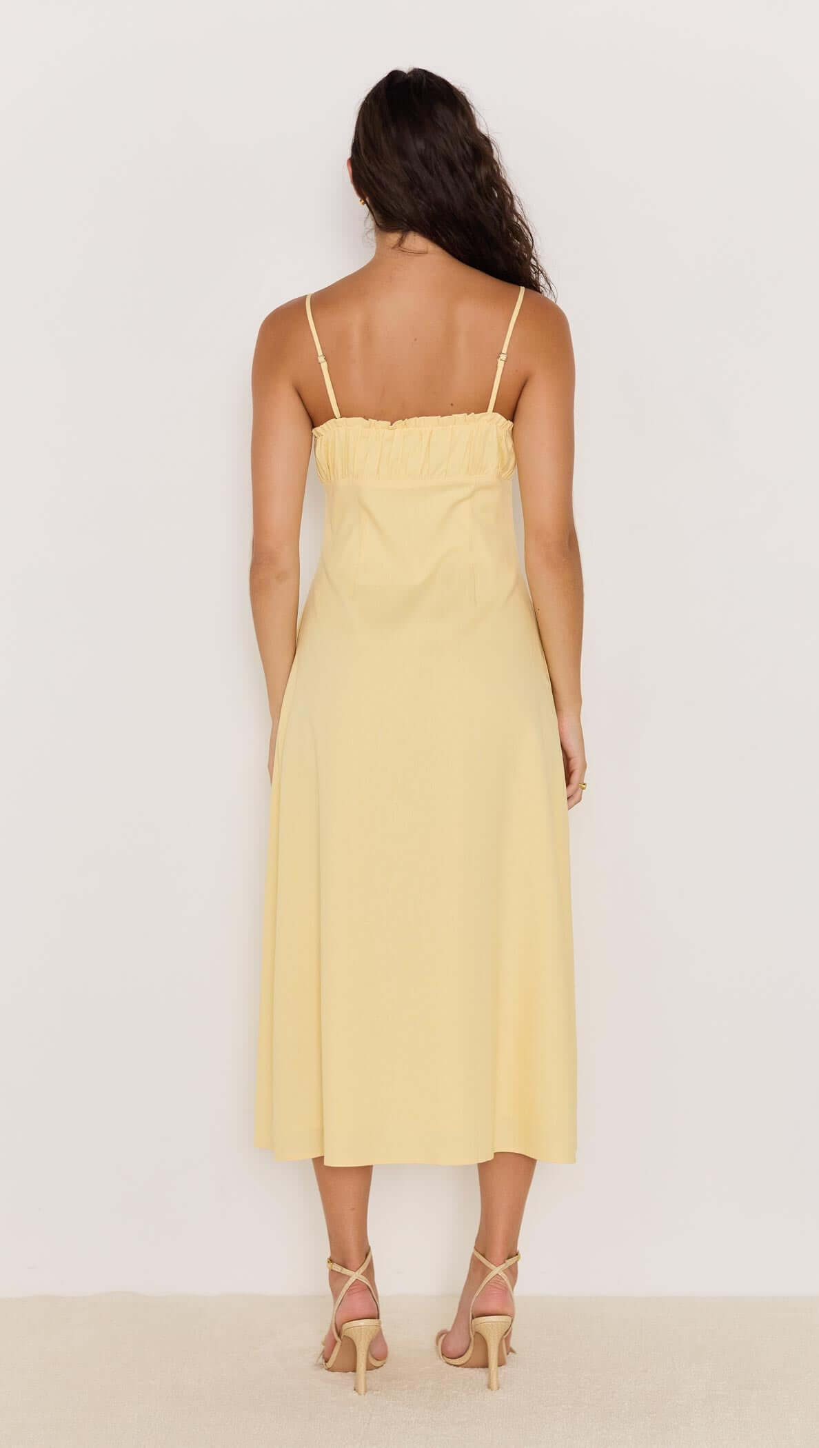 Back view of a model wearing a butter yellow bias cut midi dress with spaghetti straps.