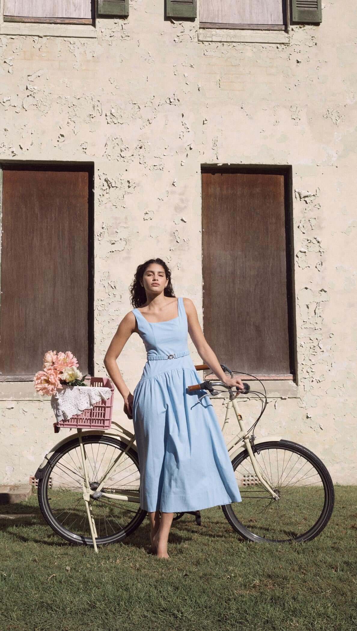 Woman in a sky blue drop waist belted midi dress posing next to a bicycle with flowers, against a vintage backdrop.